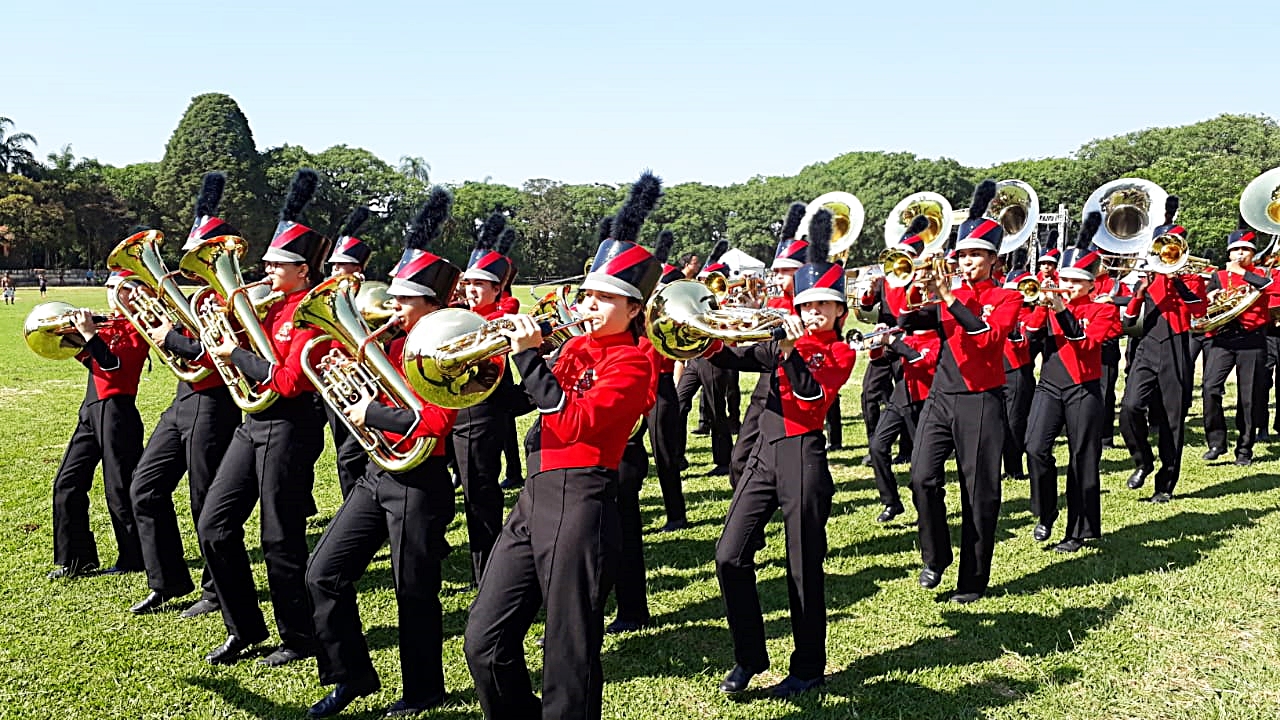 Banda Marcial de Santa Isabel tocando instrumentos musicais. Os músicos estão todos de camisa de cor vermelho, calças pretas e chapéus pretos. Eles utilizam trajes típicos de bandas marciais.